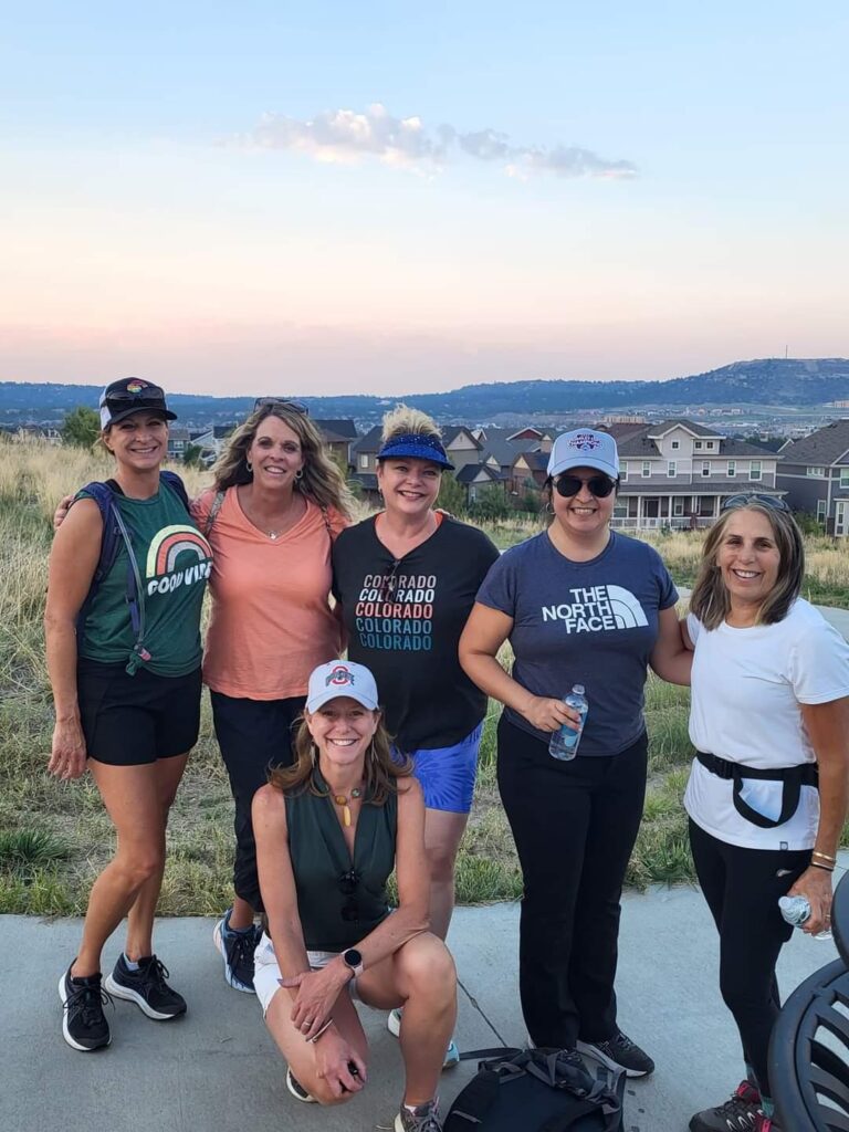 6 women who have hiked up a long hill with some houses in the background.